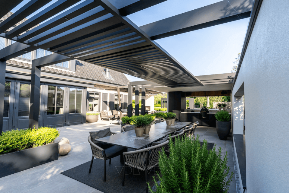 Modern buitenterras met een eettafel, stoelen en een pergola. Rondom het zitgedeelte staan ​​potplanten opgesteld. De ruimte grenst aan een moderne woning.