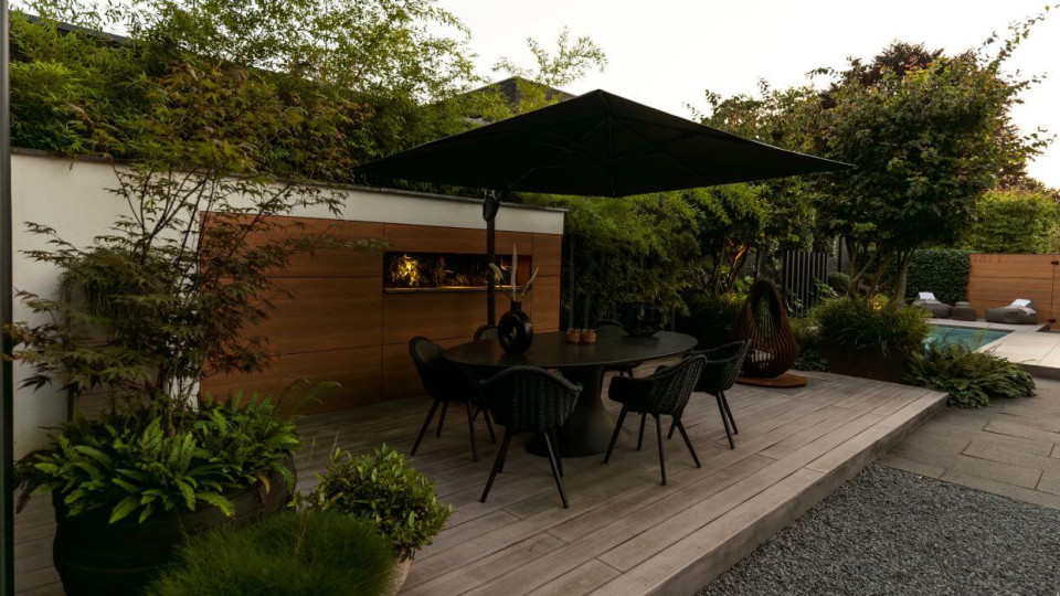 Gezellige eettafel in een industri&euml;le tuin, omringd door groenblijvende beplanting en een sfeervolle setting met pergola&rsquo;s en meerstammige bomen.