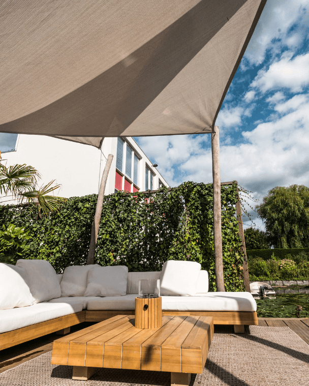 Moderne tuin met een schaduwdoek boven een loungehoek, omgeven door tropische beplanting.